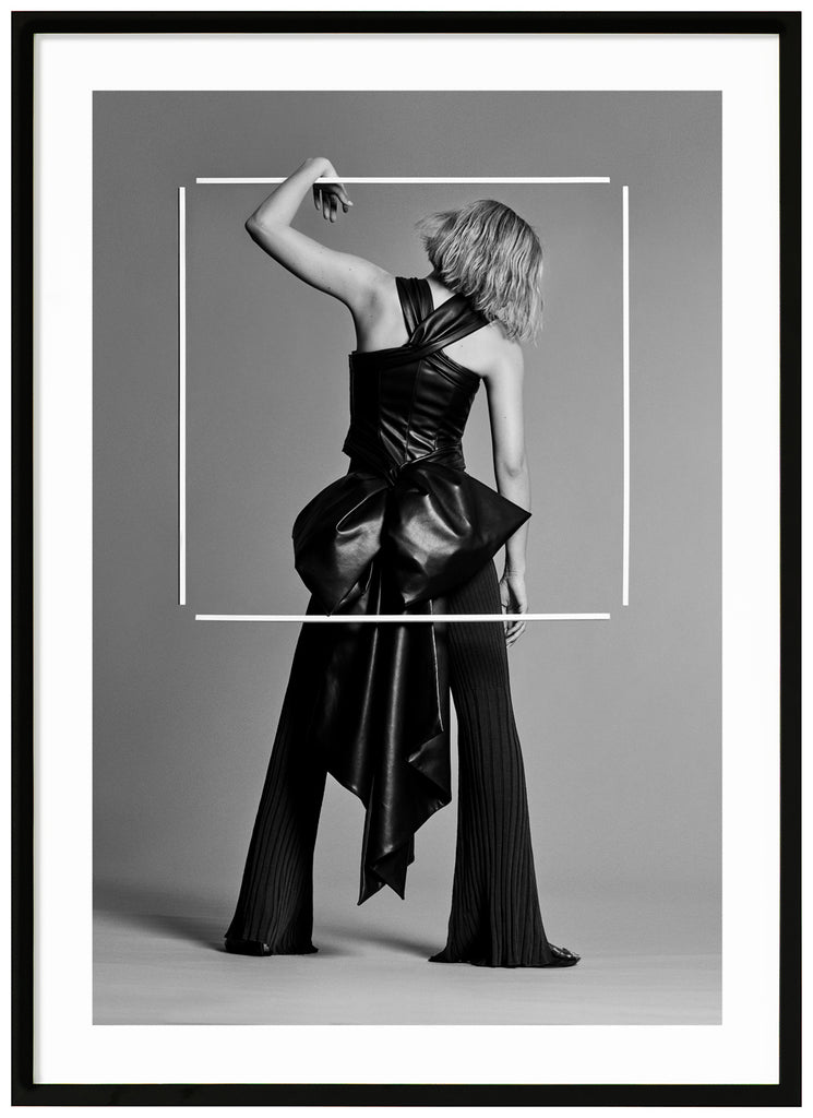 Black and white poster of woman seen from behind in photo studio. Black frame.