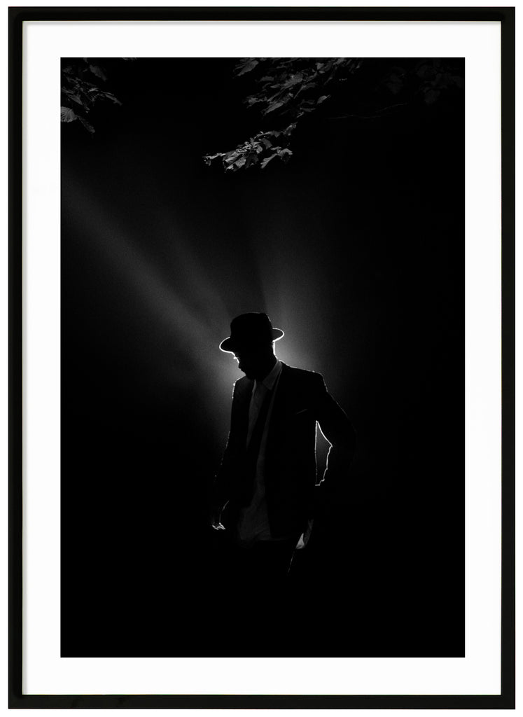 Black and white poster of a man illuminated from behind in suit and hat under some tree branches. Black frame.