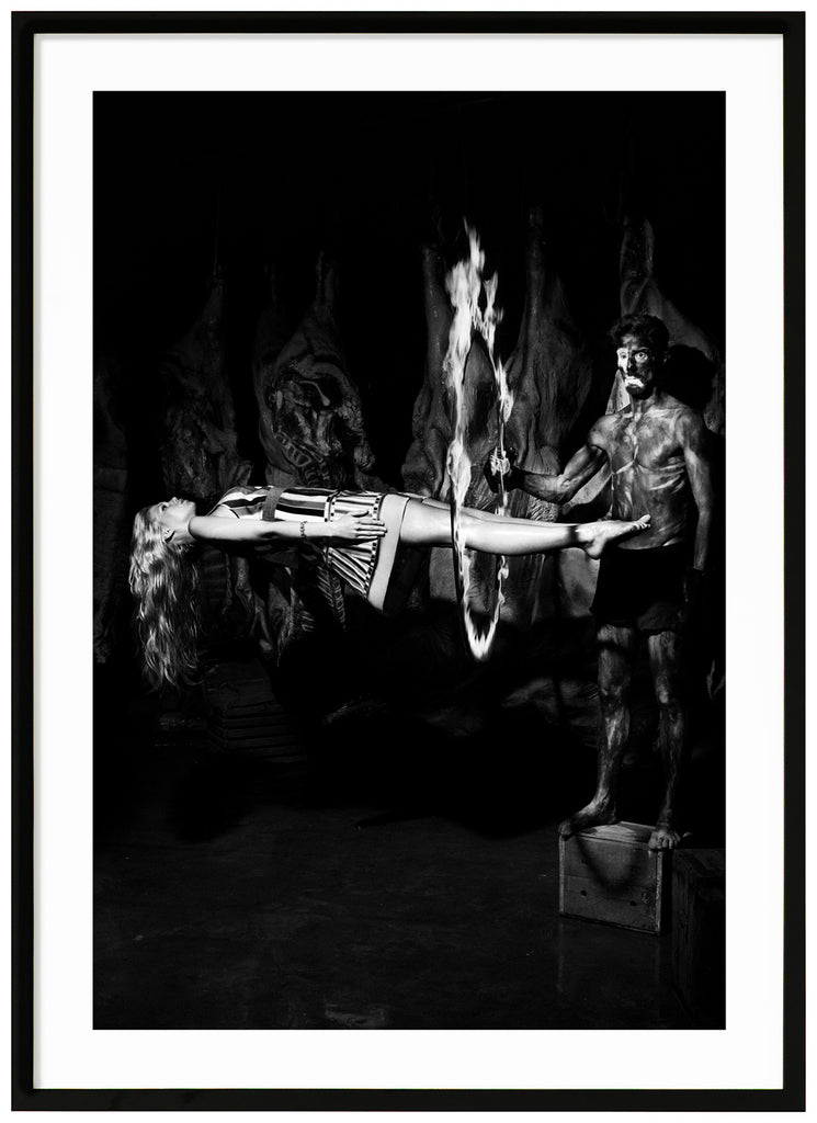 The photo shows a man standing on a wooden box holding a burning ring, with a flying woman on her back. Black frame. 