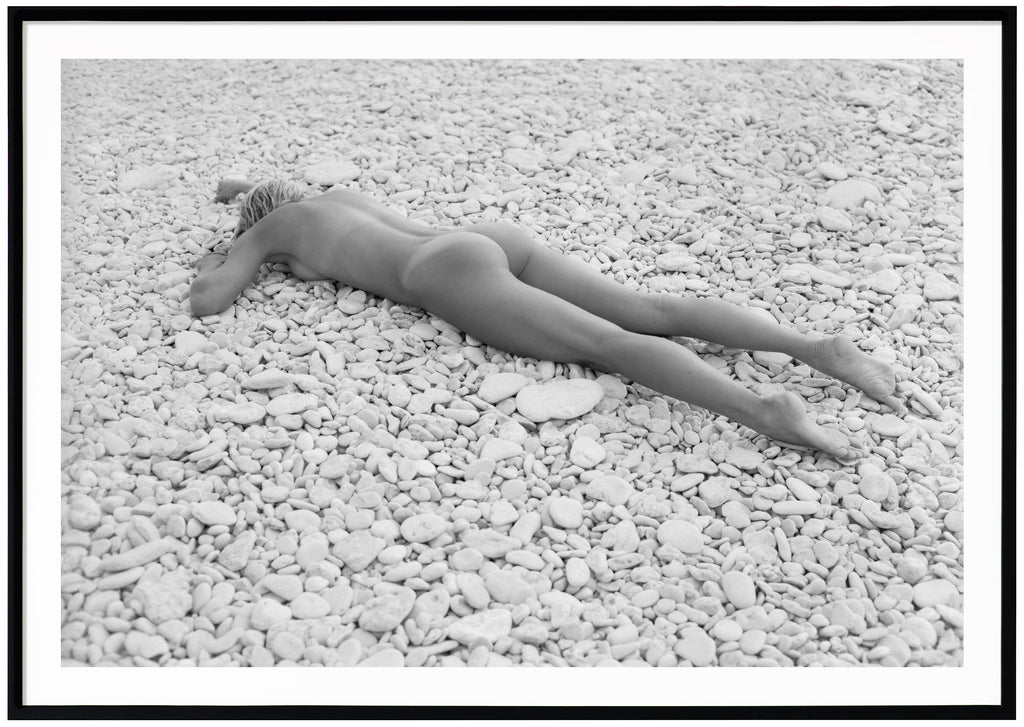 Black and white photograph of a naked woman lying on beach with white stones. Black frame. 