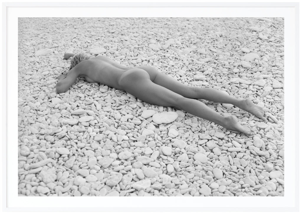 Black and white photograph of a naked woman lying on beach with white stones. White frame. 
