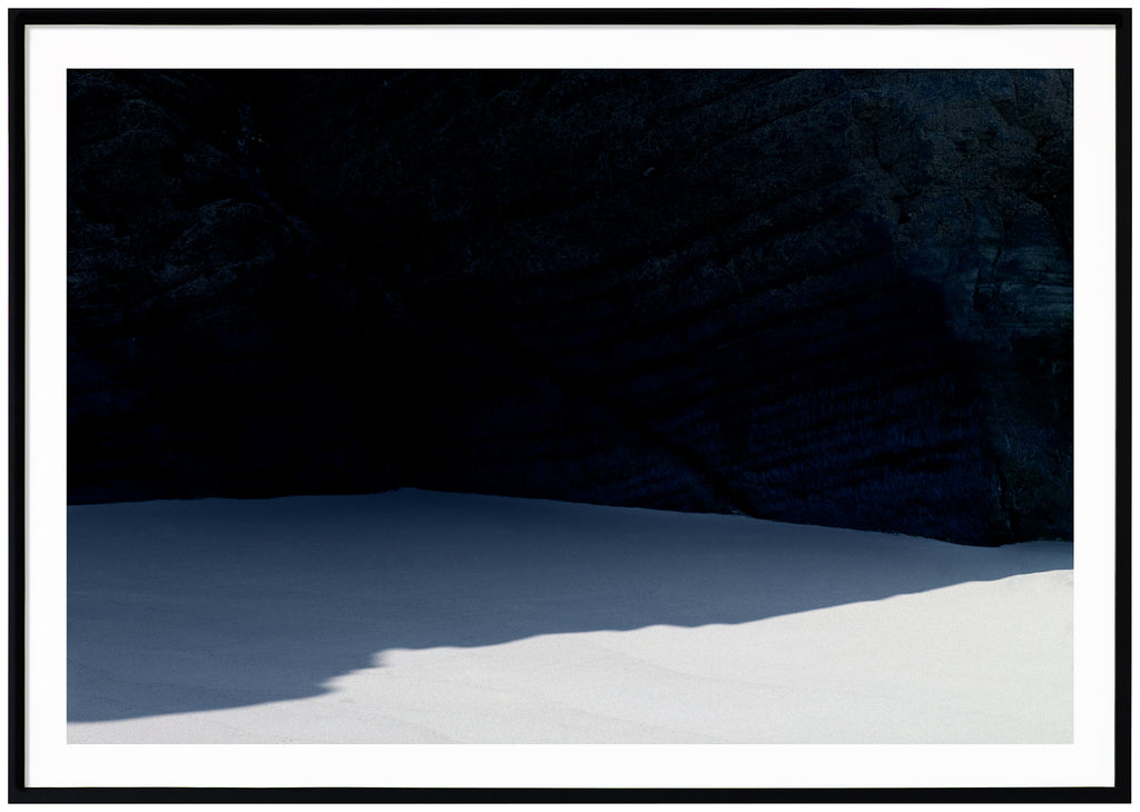  Graphic photograph of a beach with a shadow from a dark cliff.  Black frame.