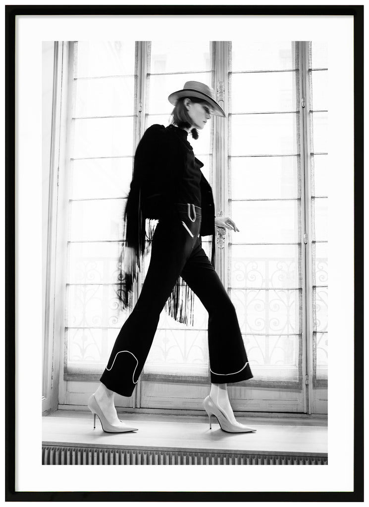 Black and white photograph of woman in front of large window seen from the side with heels hat and black dress. Black frame. 