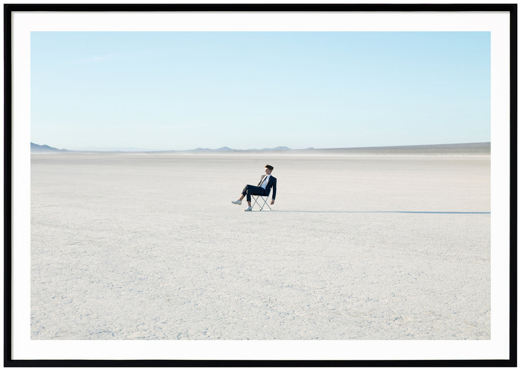 Photograph of costumed man sitting on a chair in the Mojave Desert, California.  Black frame.