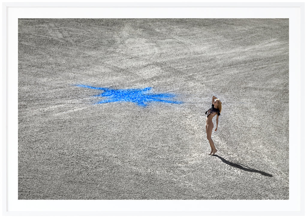Color photography in landscape format of a naked woman standing on stone ground with a blue figure on. White frame.