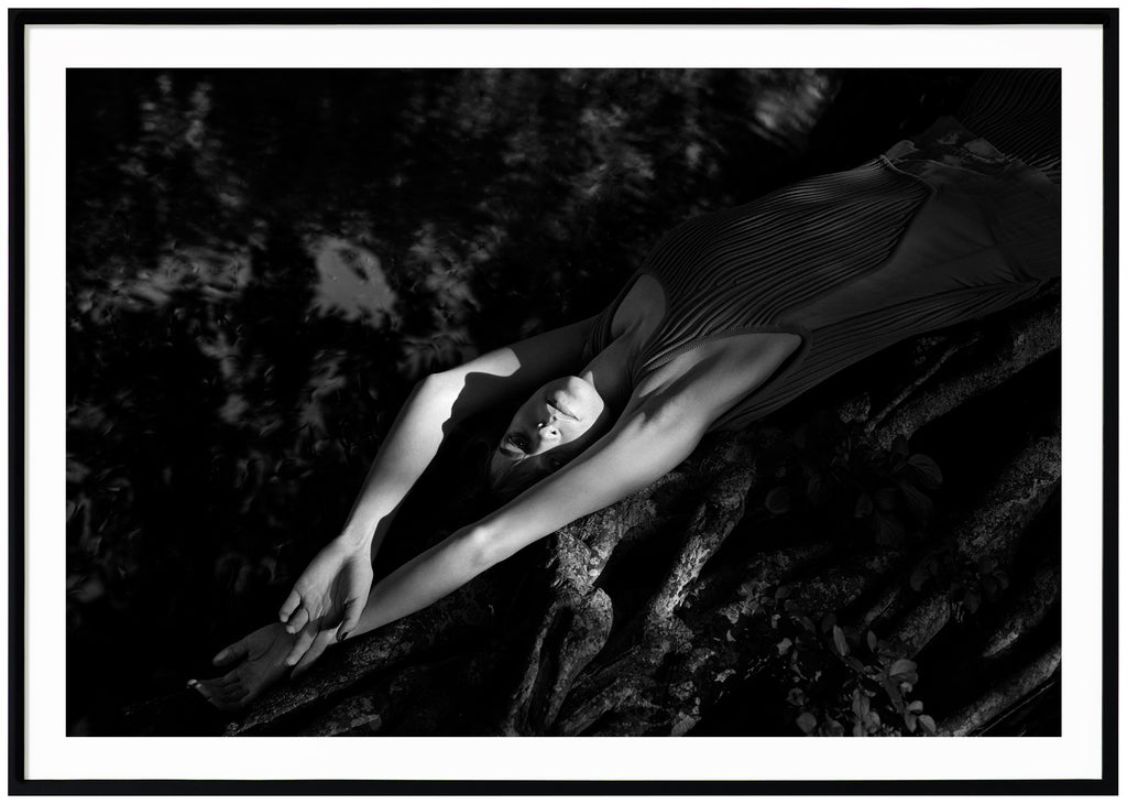Black and white photograph of a woman lying on a mangrove tree near the water. Black frame.