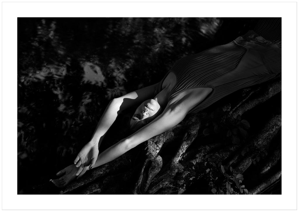 Black and white photograph of a woman lying on a mangrove tree near the water.