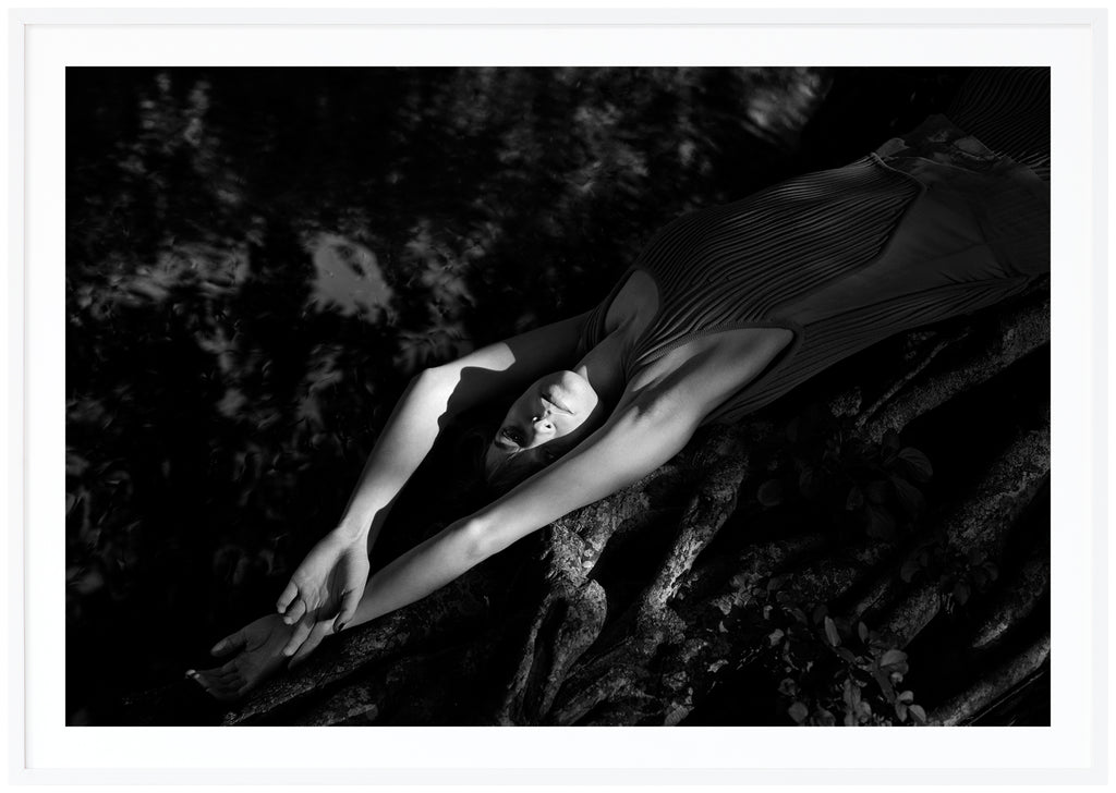 Black and white photograph of a woman lying on a mangrove tree near the water. White frame. 