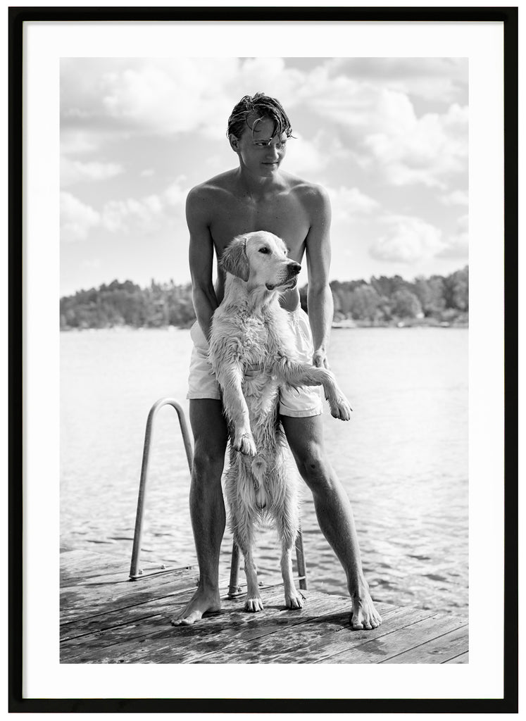 Black and white poster of guy with white dog on a jetty. A background of water and trees. Black frame. 