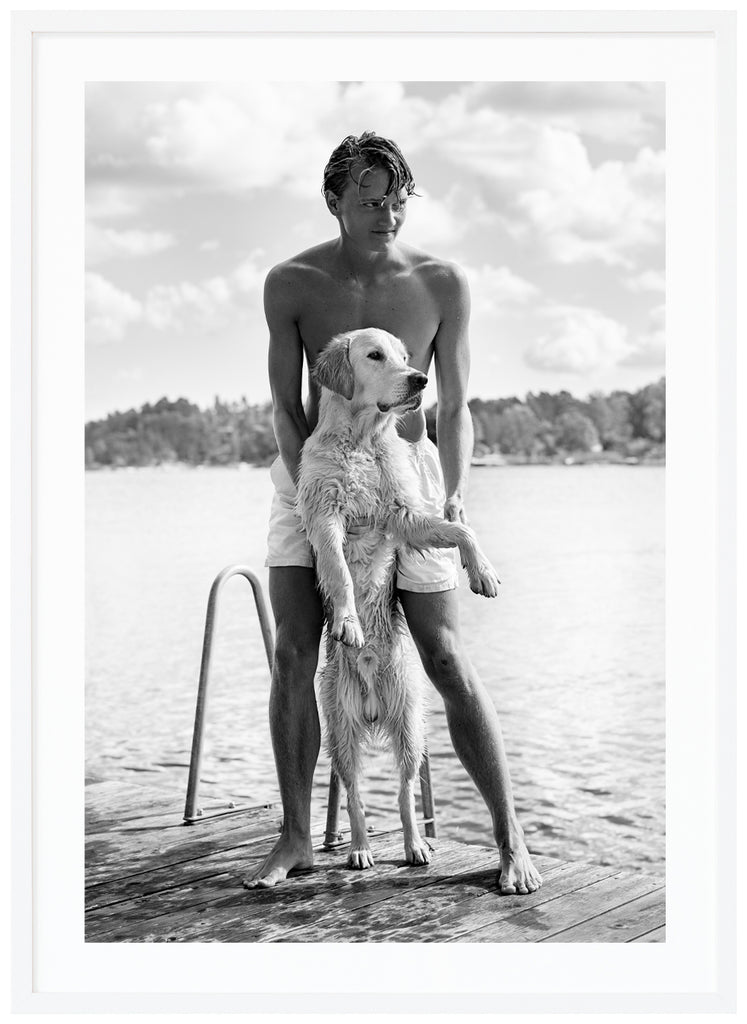 Black and white poster of guy with white dog on a jetty. A background of water and trees. White frame. 