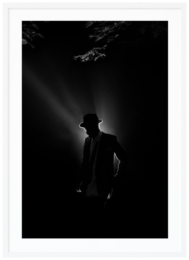 Black and white poster of a man illuminated from behind in suit and hat under some tree branches. White frame. 