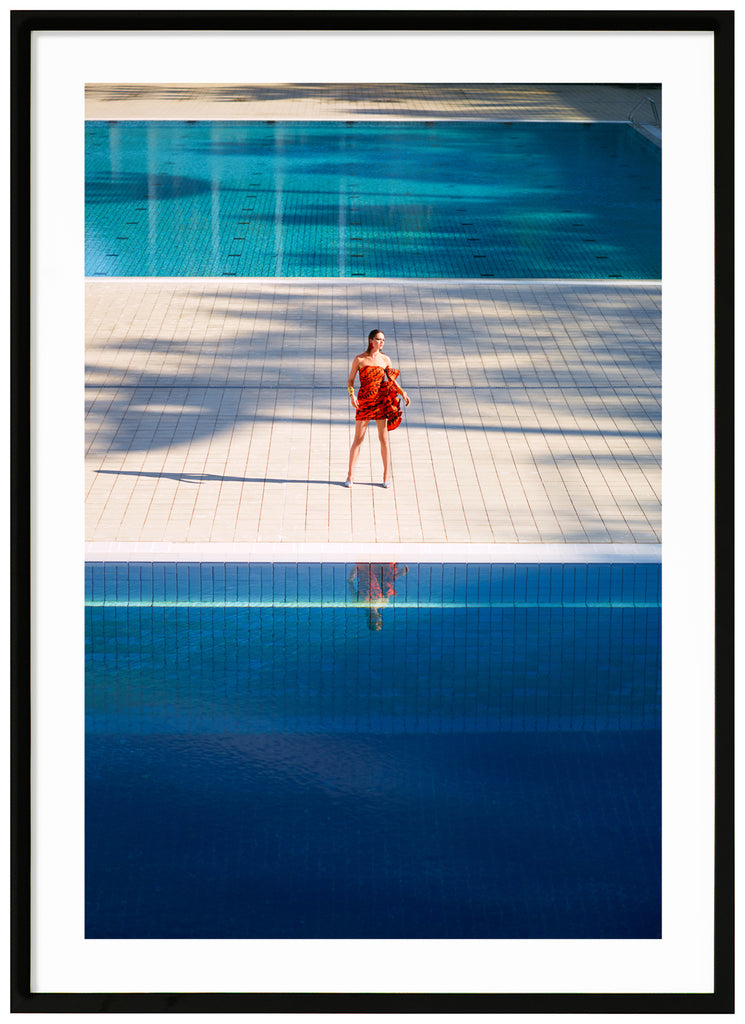 Poster of woman in orange dress standing alone between two large swimming pools. Black frame. 