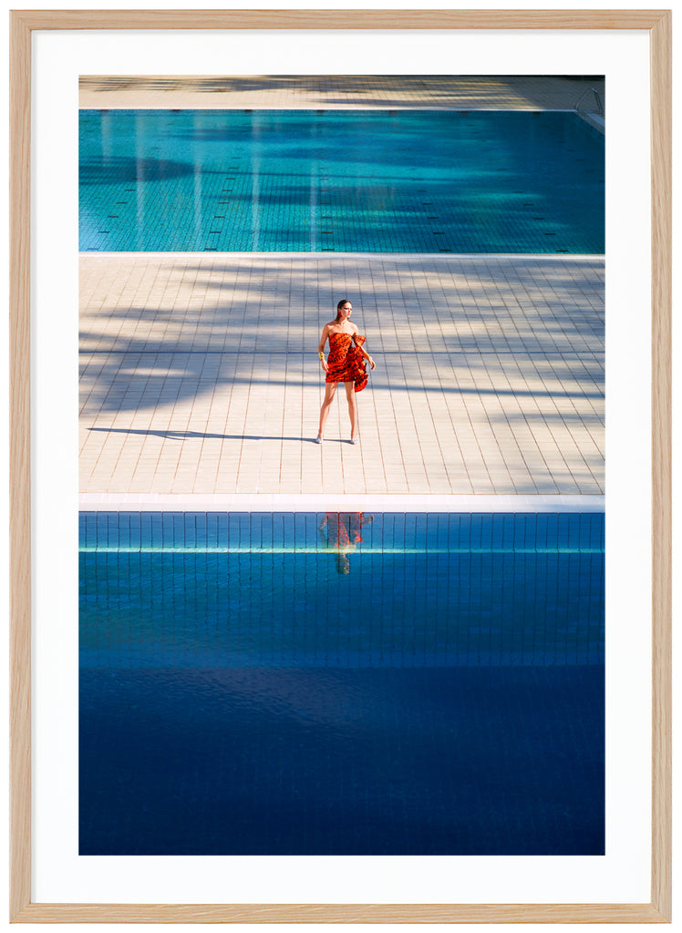 Poster of woman in orange dress standing alone between two large swimming pools. Oak frame. 
