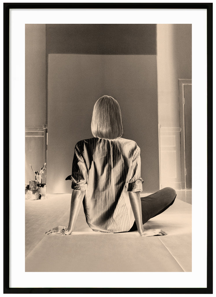 Monochrome photograph of a woman sitting in front of a blank canvas intended for painting. Black frame.