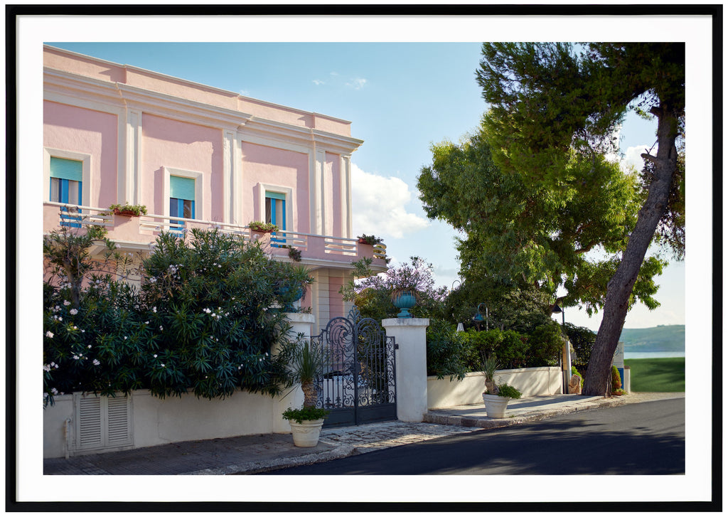Photograph of a pink house with blue window frames and turquoise shutters located in Italy. Black frame.