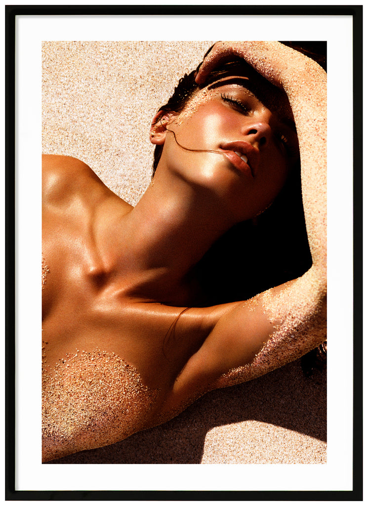 Photograph of a posed woman lying on her back with sand on her body. Black frame. 