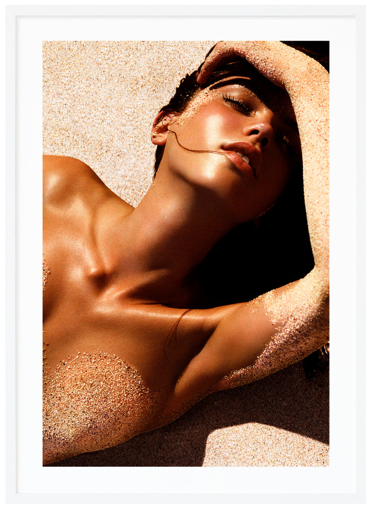 Photograph of a posed woman lying on her back with sand on her body.  White frame. 