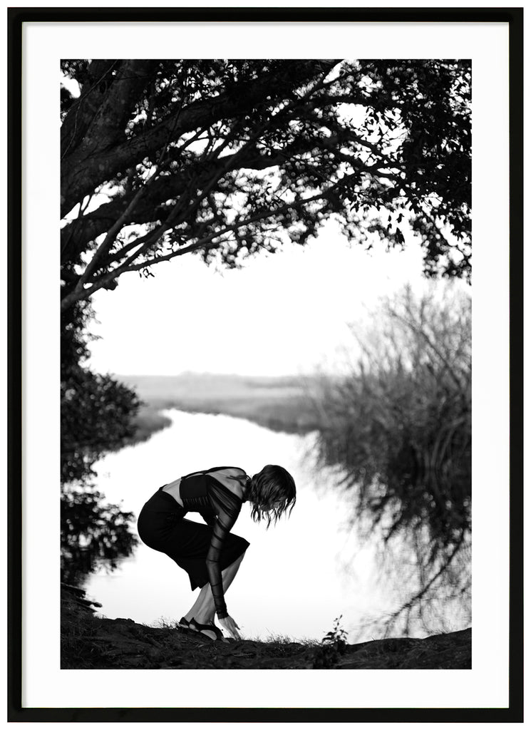 Black and white photograph of a woman under a tree with water in the background. Black frame.