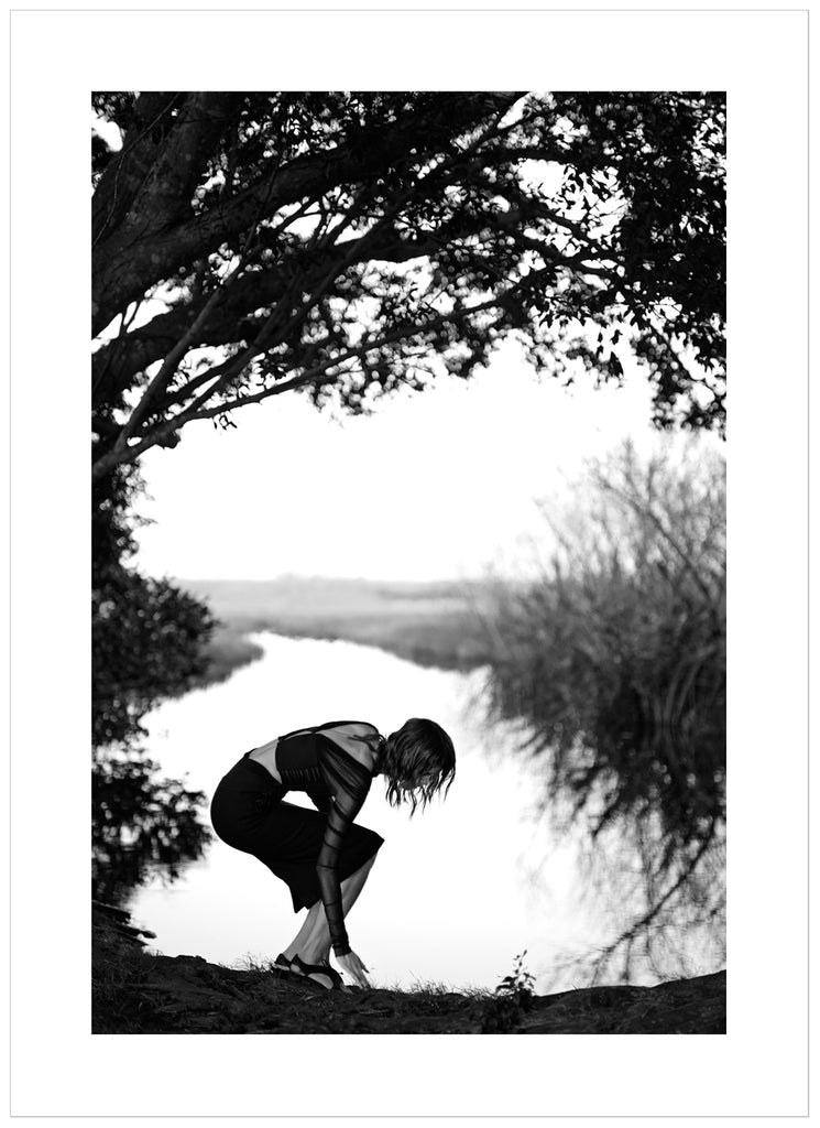 Black and white photograph of a woman under a tree with water in the background. 