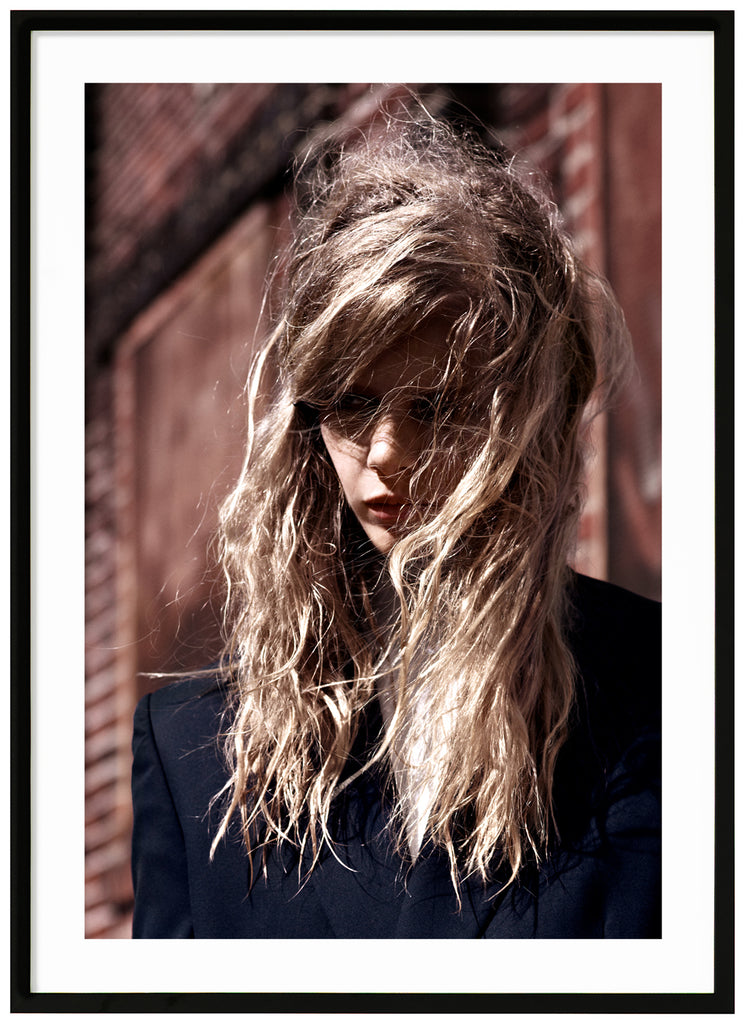 Color photograph of a woman, whose hair blows lightly over her face as she looks down. Black frame. 