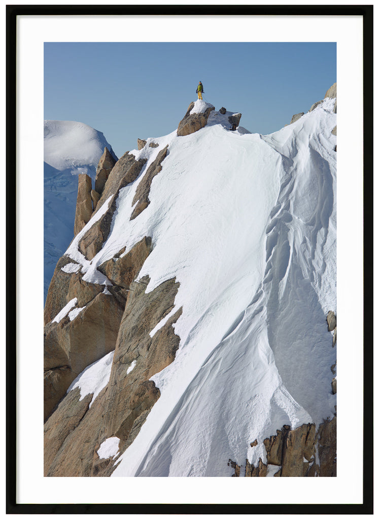 Color photograph of a lone climber in yellow pants and green jacket, standing on top of a snow-capped cliff.  Black frame. 