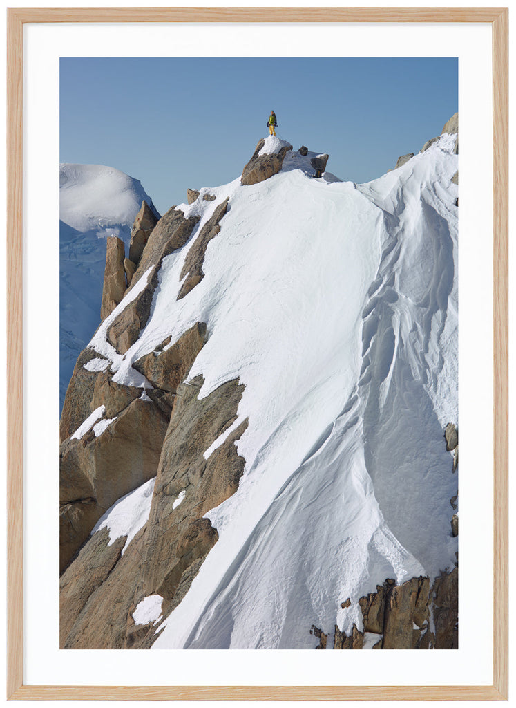 Color photograph of a lone climber in yellow pants and green jacket, standing on top of a snow-capped cliff.  Oak frame. 