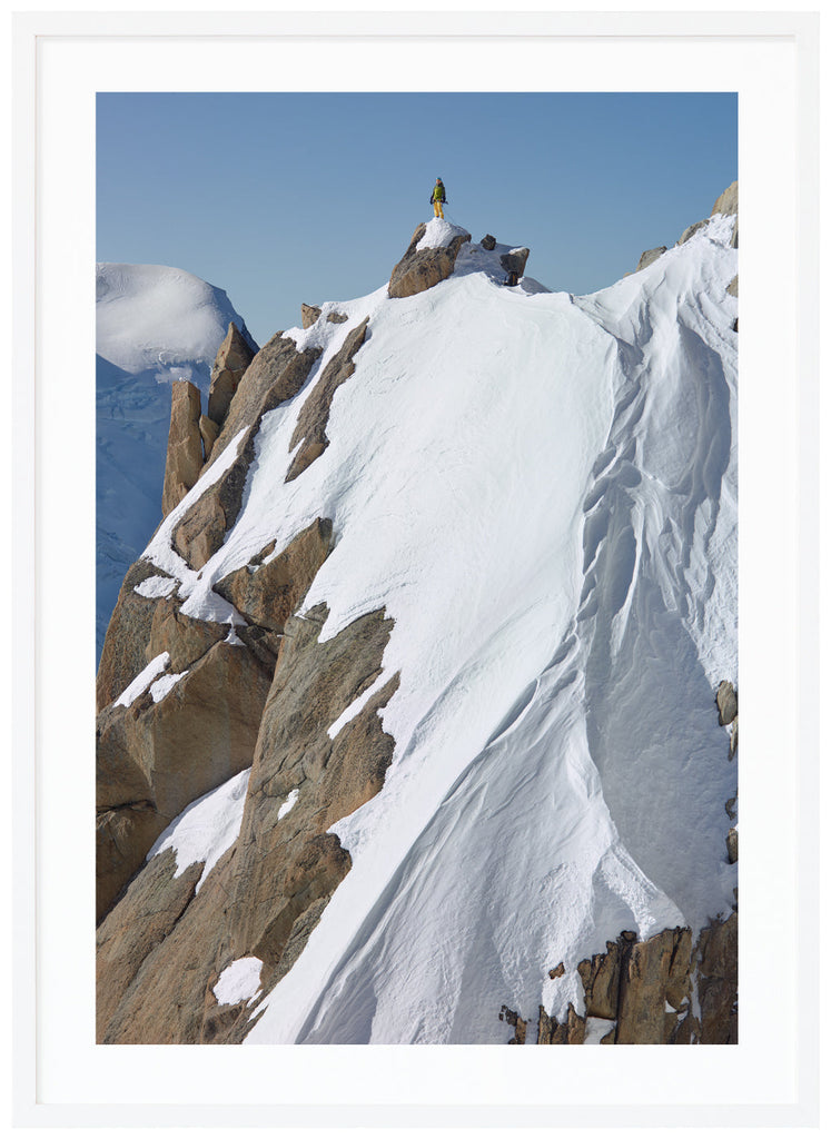 Color photograph of a lone climber in yellow pants and green jacket, standing on top of a snow-capped cliff.  White frame. 