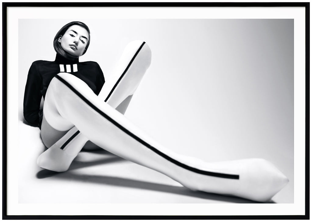 Black and white photograph of woman sitting on the floor. White overknee heels with black stripe. Black shirt. Black frame. 