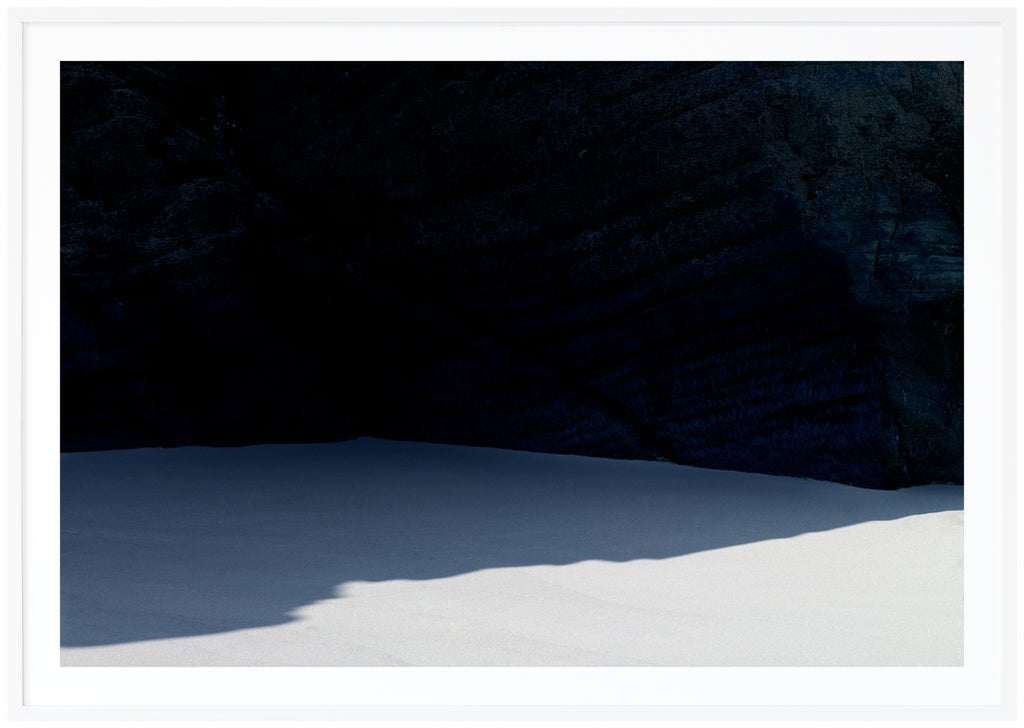  Graphic photograph of a beach with a shadow from a dark cliff.  White frame. 