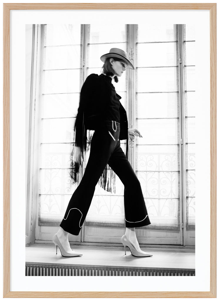 Black and white photograph of woman in front of large window seen from the side with heels hat and black dress. Oak frame.