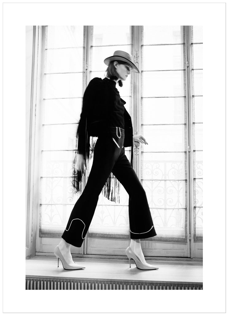 Black and white photograph of woman in front of large window seen from the side with heels hat and black dress.