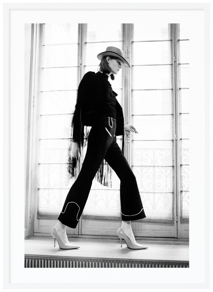 Black and white photograph of woman in front of large window seen from the side with heels hat and black dress. White frame.