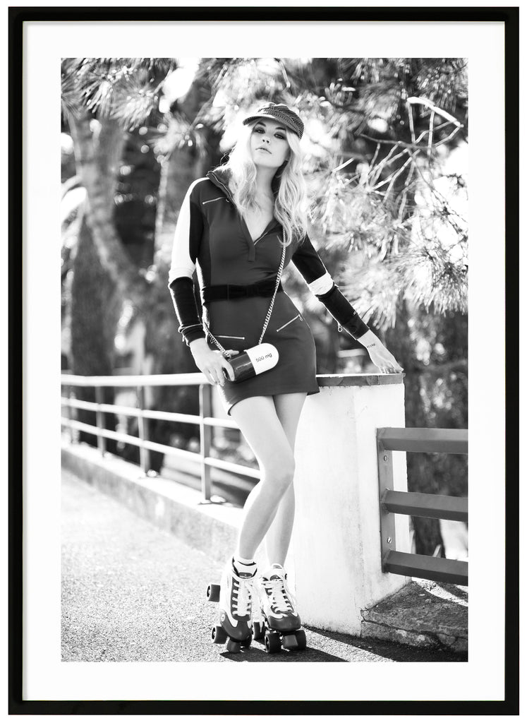 Black and white photograph of woman with roller skates on her feet by a fence, with trees in the background. Black frame. 