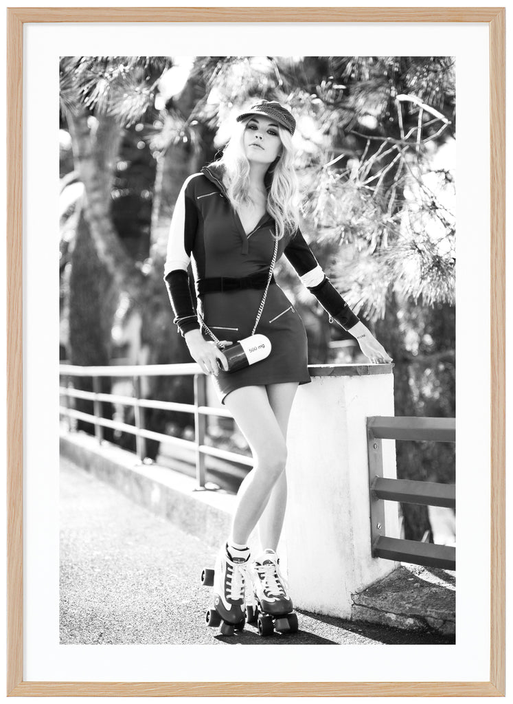 Black and white photograph of woman with roller skates on her feet by a fence, with trees in the background. Oak frame.