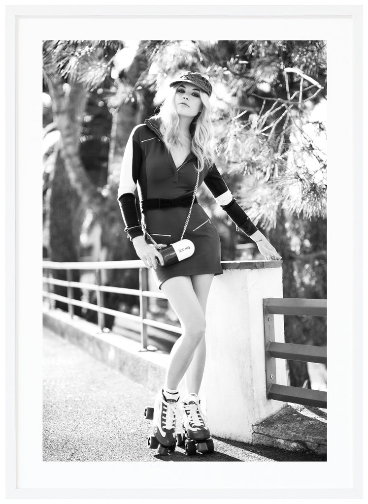 Black and white photograph of woman with roller skates on her feet by a fence, with trees in the background. White frame. 