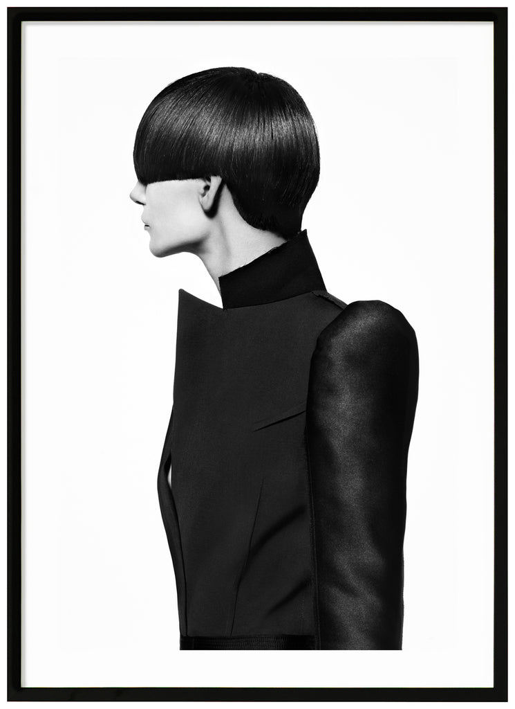 Black-and-white photograph of a female model with a bob-like hairstyle and wearing a black coat. Black frame. 