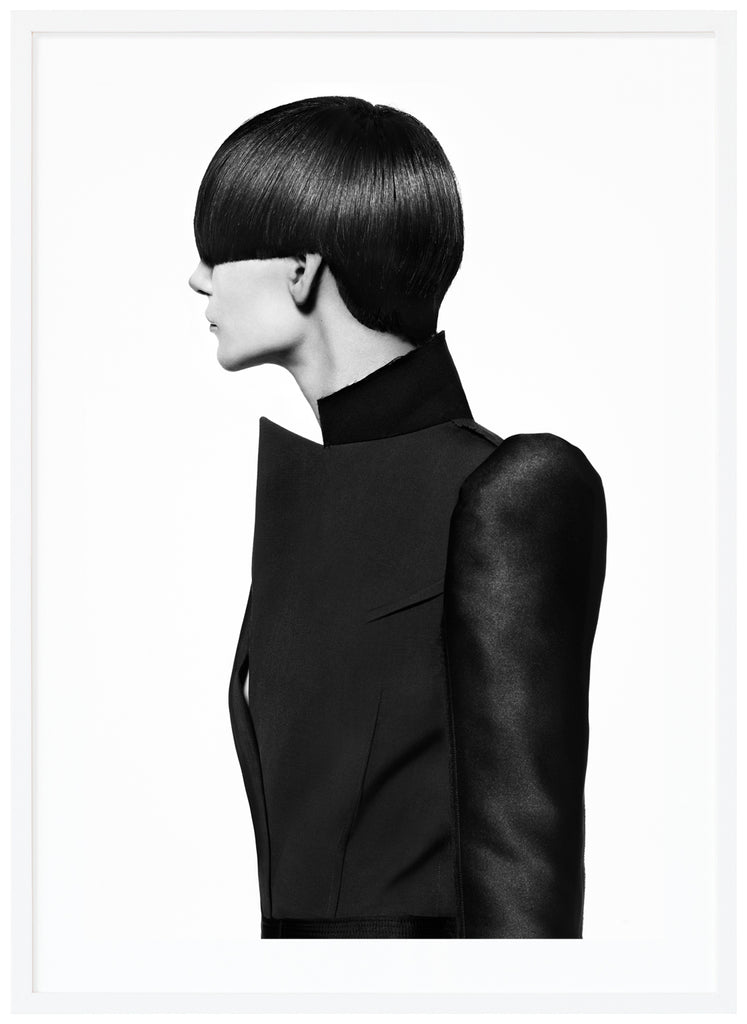 Black-and-white photograph of a female model with a bob-like hairstyle and wearing a black coat. White frame. 