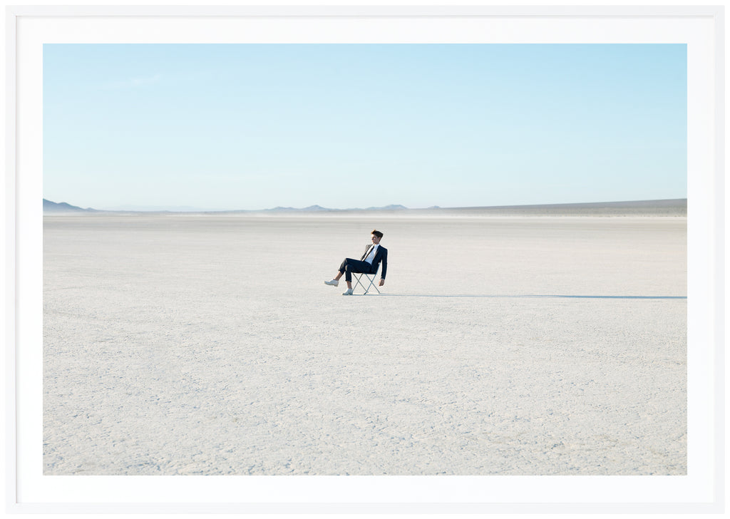 Photograph of costumed man sitting on a chair in the Mojave Desert, California.  White frame. 