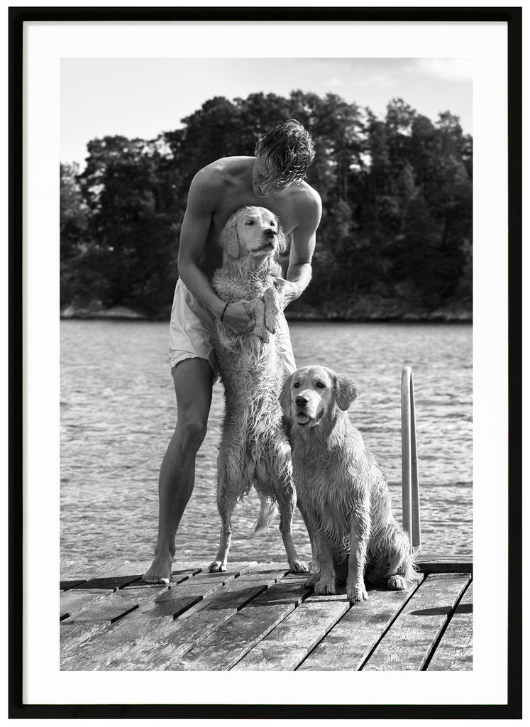 Black and white poster of guy with dogs on a wet jetty. Portrait format. Black frame. 