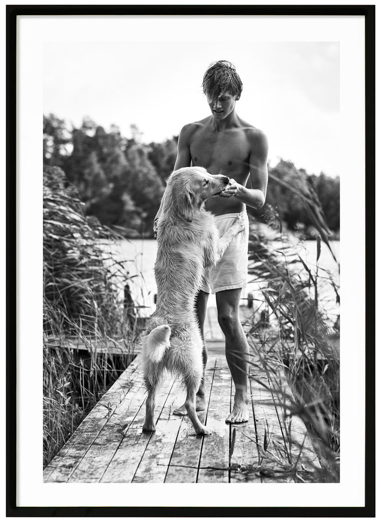 Black and white poster of guy in white shorts with white dog in front of him. On the jetty among reeds. Water and trees in the background. Black frame. 