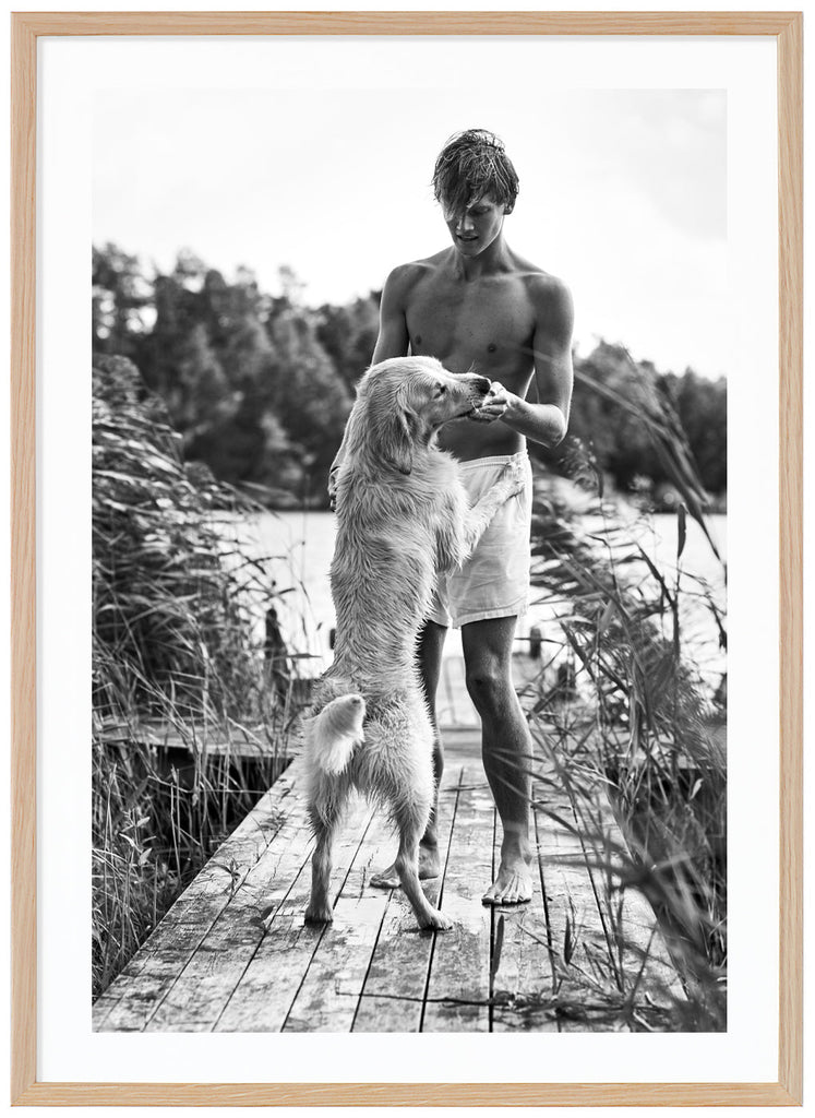 Black and white poster of guy in white shorts with white dog in front of him. On the jetty among reeds. Water and trees in the background. Oak frame. 