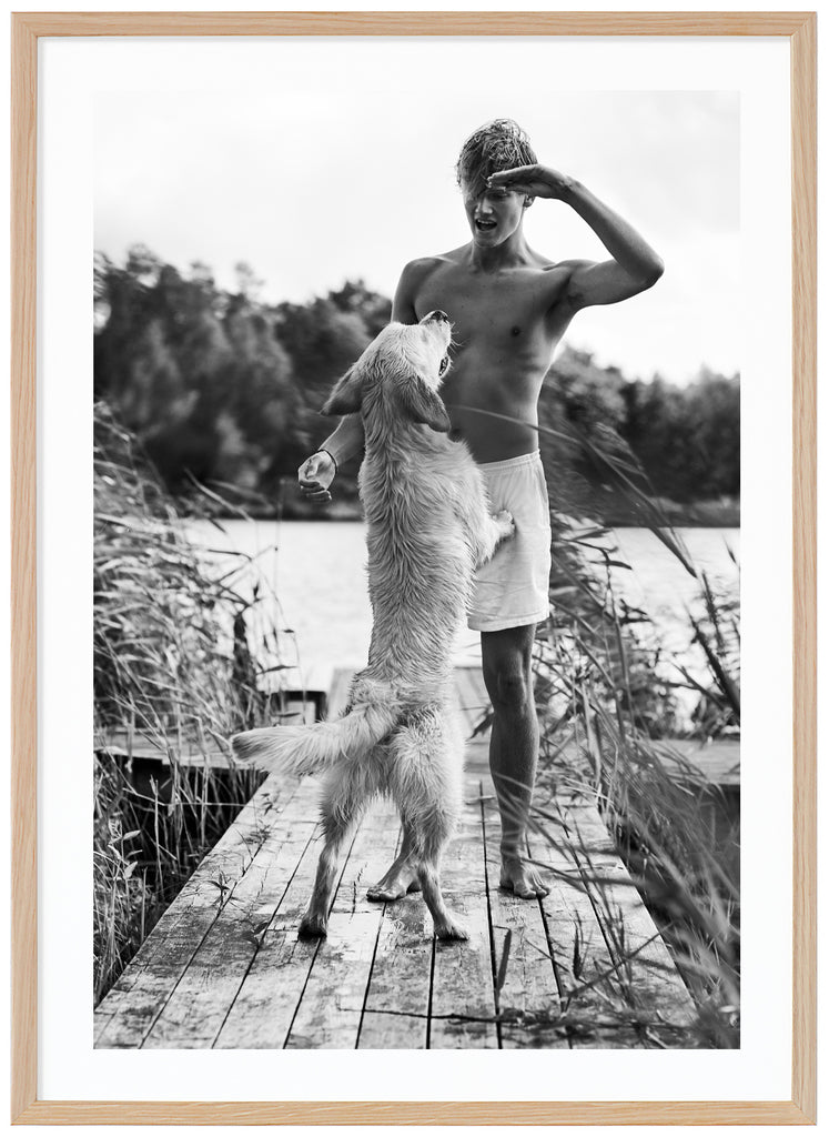 Black and white poster of guy in topless playing with white dog on jetty among reeds. Oak frame. 