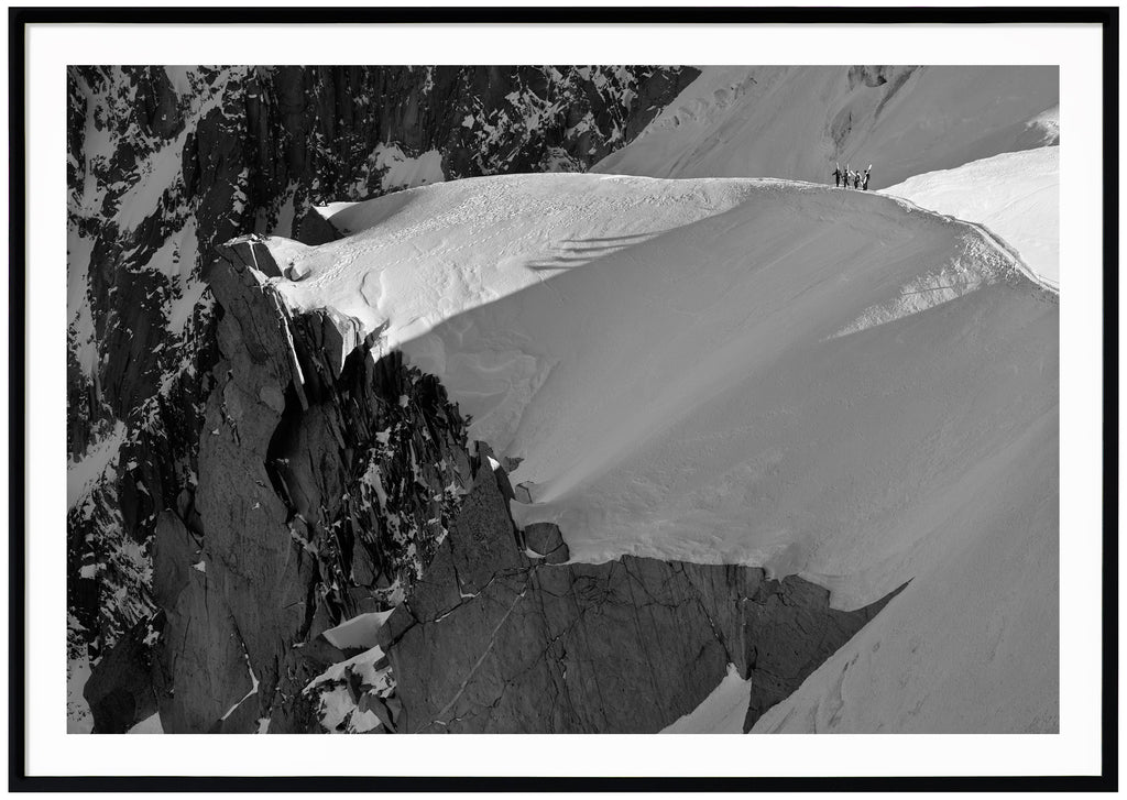 Black and white photograph of four skiers hiking on a snow-capped ridge. Landscape format. Black frame. 
