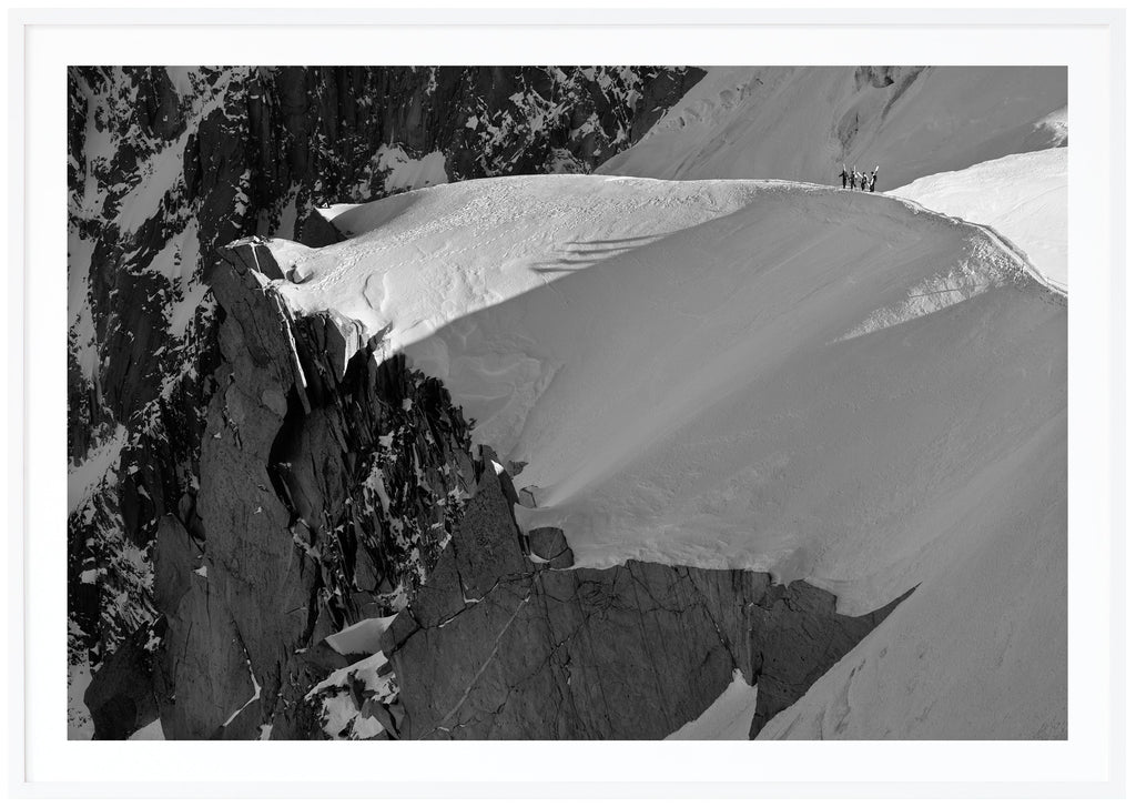 Black and white photograph of four skiers hiking on a snow-capped ridge. Landscape format. White frame. 