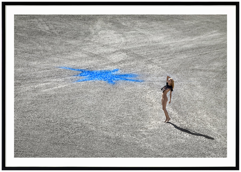Color photography in landscape format of a naked woman standing on stone ground with a blue figure on. Black frame.