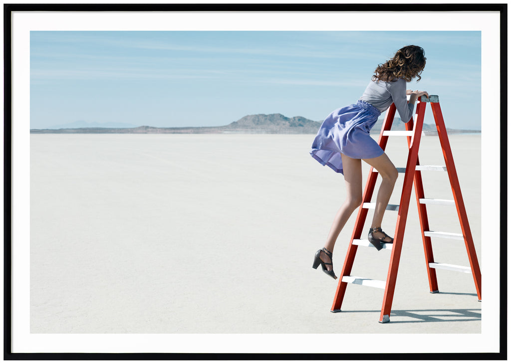 Poster of woman climbing on orange ladder in the desert. Horizontal format. Black frame.