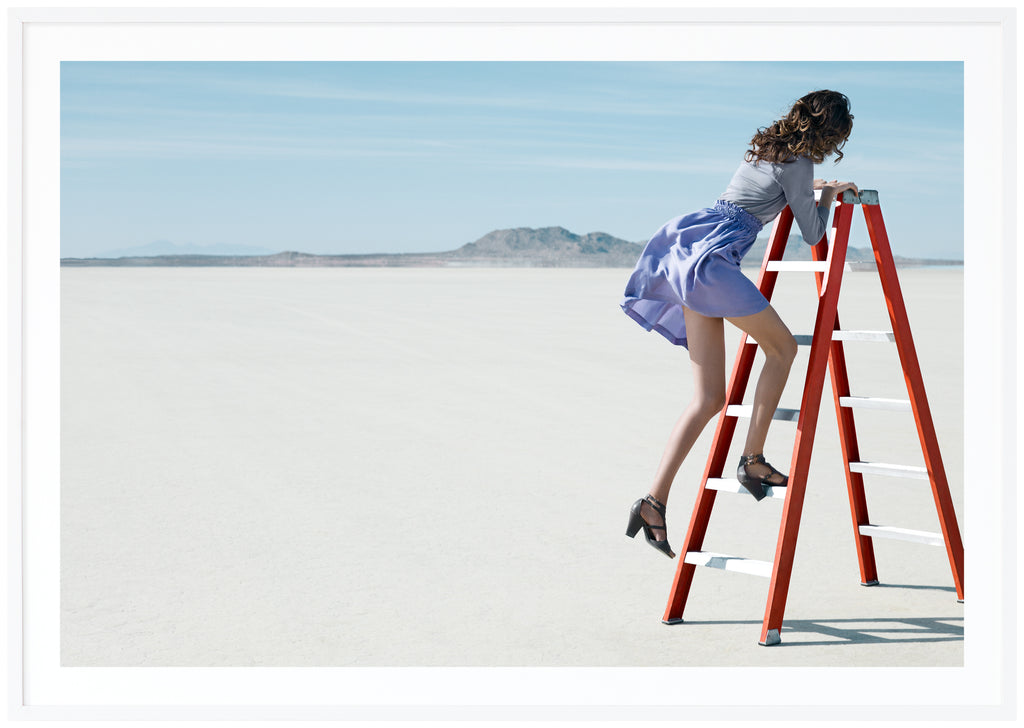 Poster of woman climbing on orange ladder in the desert. Horizontal format. White frame. 