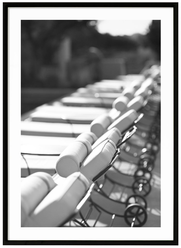 Black and white photograph of several lined up white sun loungers in the sun. Black frame. 