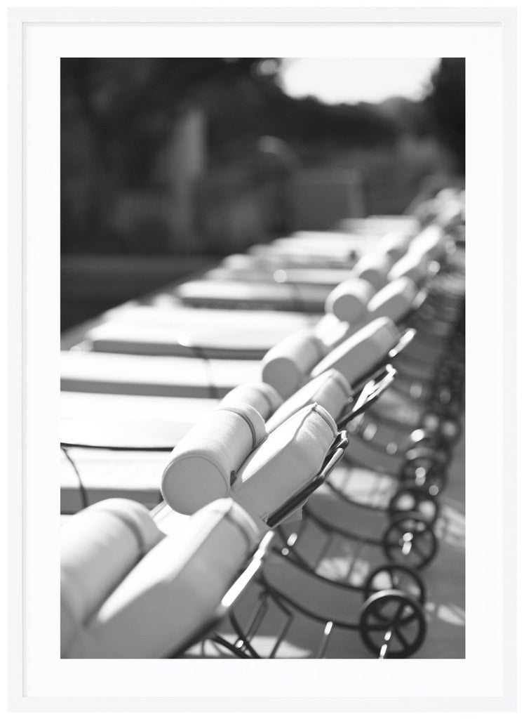 Black and white photograph of several lined up white sun loungers in the sun. White frame. 