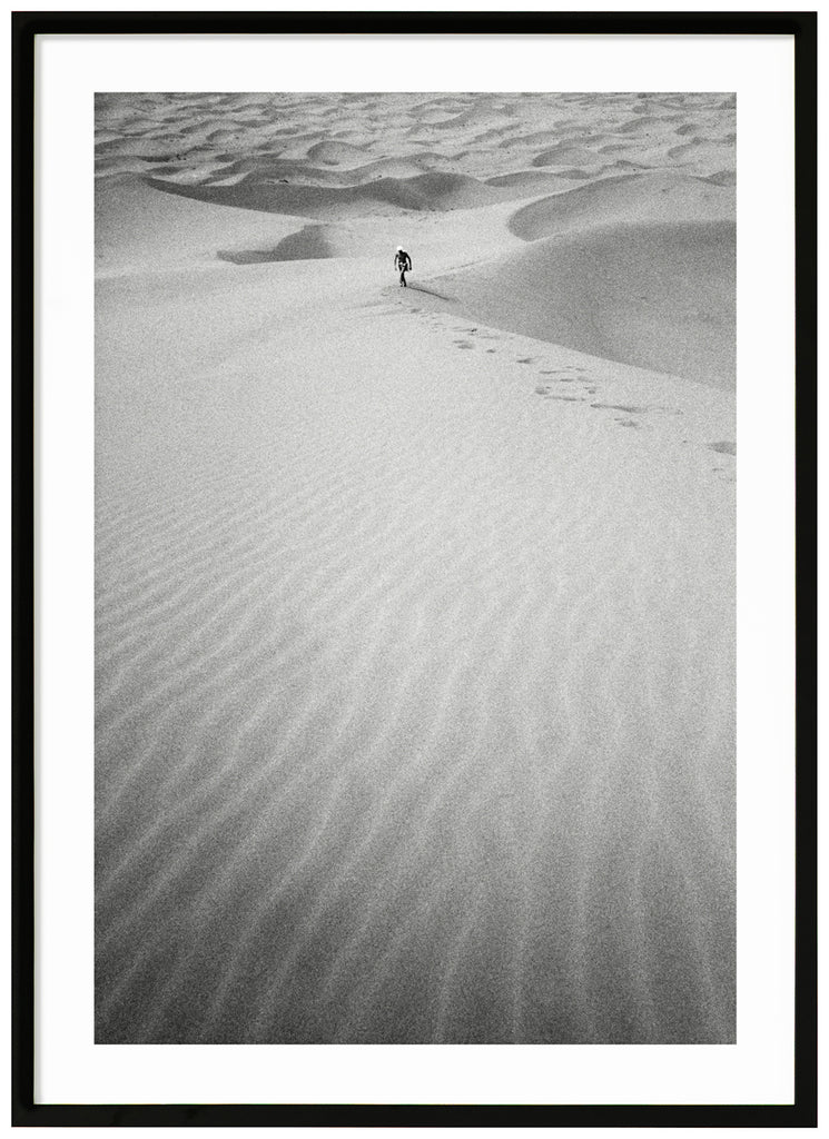 Black and white analog photograph of a man in the distance, walking up a huge sand dune in the middle of the Sahara desert. Black frame. 